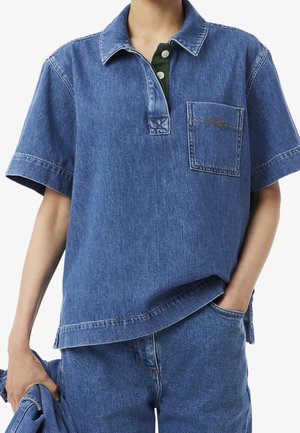 Osoba nosząca krótkim rękawem niebieskie dżinsowe polo z kieszenią na piersi na zielonej koszuli z kołnierzykiem, trzymająca dopasowaną niebieską dżinsową kurtkę.