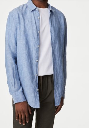 Man wearing a light blue linen button-up shirt over a white t-shirt and dark pants, standing against a plain white background.