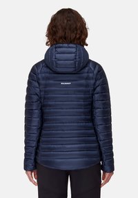 Person med lockigt brunt hår som bär en marinblå vadderad jacka med huva och "Mammut"-logotyp på ryggen, stående mot en enfärgad bakgrund.