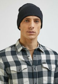 Czarna, ribbed dziana czapka na głowie modela, w połączeniu z czarno-białą koszulą flanelową w kratę z zapięciem na guziki.