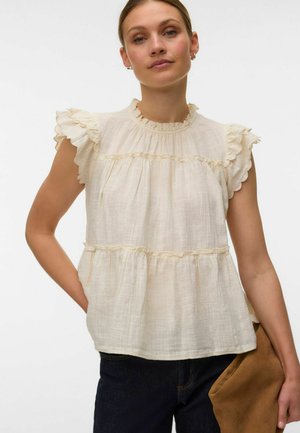 Frau trägt eine cremefarbene, ärmellose Rüschenbluse und dunkle Hose und hält eine braune Clutch vor einem weißen Hintergrund.