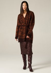 Cardigan marrone e arancione con motivo, aperto davanti con un nodo. Abbinato a una gonna di paillettes bordeaux scintillante e stivali bordeaux fino al ginocchio.