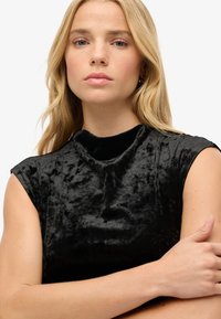 Czarna aksamitna bluzka o wysokim, okrągłym dekolcie i rękawach typu cap, z delikatnie teksturowaną powierzchnią. Gładkie i miękkie wykończenie.