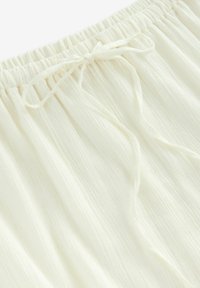 Tessuto bianco e texturizzato con vita elasticizzata e dettaglio con cordino annodato. Il tessuto sembra leggero e leggermente stropicciato.