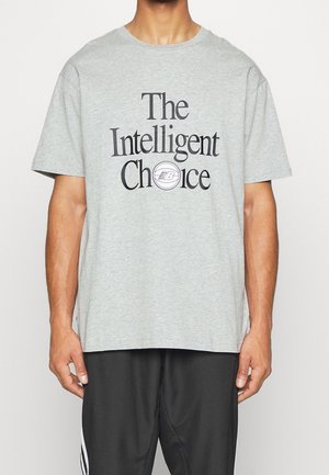 Person trägt ein hellgraues T-Shirt mit dem Schriftzug „The Intelligent Choice“ und dem New Balance-Logo, kombiniert mit schwarzen Sporthosen.