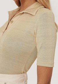 Polo à manches courtes en tissu côtelé beige clair avec col et détail d'un bouton au niveau du cou, associé à un pantalon de couleur claire.
