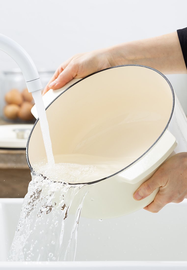 Cremfarbener Schüssel mit einem dunklen Rand, die von einem weißen Wasserhahn gefüllt wird, der von einer Hand gehalten wird. Die Schüssel hat zwei seitliche Griffe.