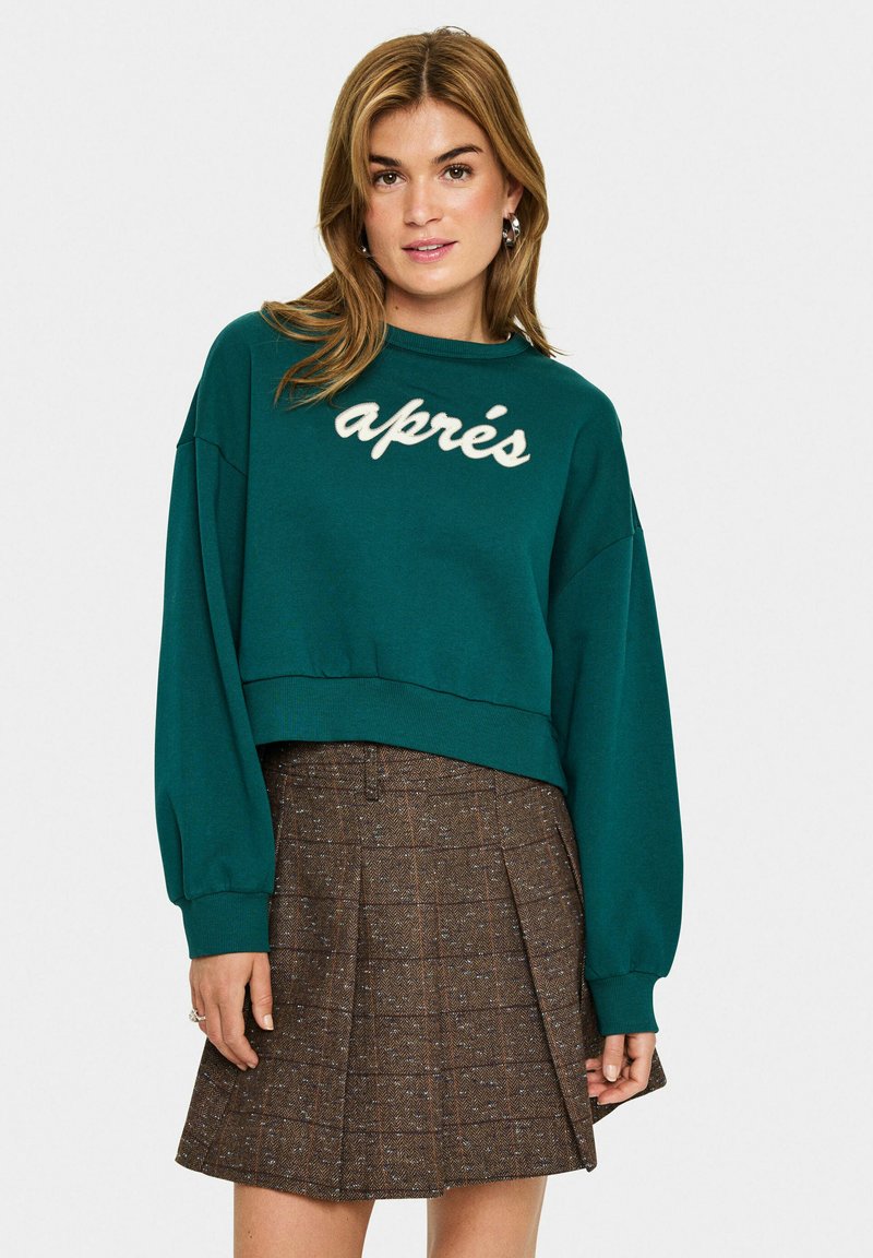 Teal cropped sweatshirt met witte "après" opdruk, met verlaagde schouders en geribde zoom, gecombineerd met een bruine ruitenrok.