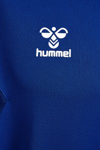 Maglietta sportiva blu realizzata in tessuto testurizzato e traspirante. Presenta il logo "hummel" bianco e un design grafico sulla parte superiore sinistra.