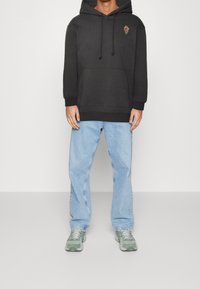 Felpa nera con tasca frontale, cordoncini e ricamo di rose. Indossata con jeans azzurri e sneaker verdi su uno sfondo neutro.