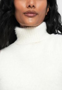 Frau mit mittlerem Hautton, langen schwarzen Haaren, trägt einen weißen, flauschigen Rollkragenpullover, Lippen geschlossen, neutrale Miene.