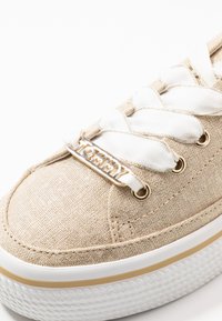 Sneaker in tessuto beige con lacci bianchi, occhielli dorati e logo con dettagli dorati. Parte superiore testurizzata e suola in gomma bianca con scanalature.