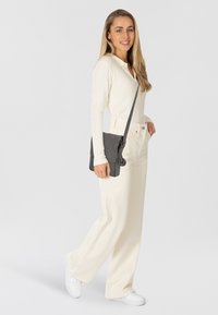 Maglia a maniche lunghe leggera con righe, pantaloni beige e sneakers bianche. Borsa a tracolla nera con dettaglio a forma di ancora. Modella presenta un outfit casual.