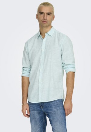 Giovane uomo con capelli biondi corti che indossa una camicia azzurro chiaro a maniche lunghe con le maniche arrotolate e jeans blu, in piedi davanti a uno sfondo semplice.