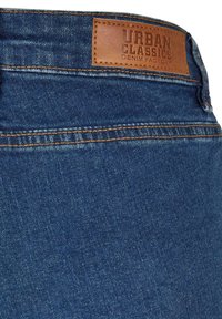 Gros plan sur un jean en denim bleu montrant la ceinture arrière avec des coutures orange et une étiquette en cuir marron portant l'inscription "Urban Classics Denim Factory."