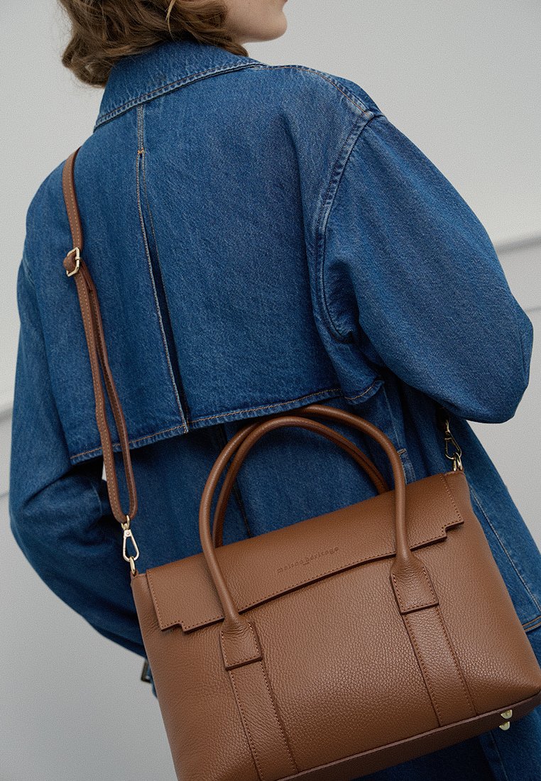 Braune Lederhandtasche mit zwei oberen Griffen und einem abnehmbaren Schulterriemen, mit einer strukturierten Oberfläche und einem kantigen Klappenverschluss, getragen über einer Jeansjacke.