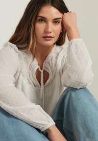 Blusa bianca trasparente con pois in rilievo, scollatura volants e maniche a sbuffo. Abbinata a jeans in denim azzurro chiaro.