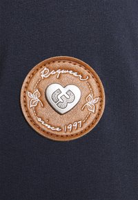 Brun läderlapp med präglat silverhjärta och sammanflätade initialer, sydd på mörkt tyg, med texten "Raquwear sedan 1997".