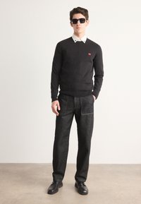 Pulover negru cu manșete cu nervuri, cămașă albă cu guler dedesubt, pantaloni din denim închis și pantofi negri, accesorizat cu ochelari de soare închiși la culoare.