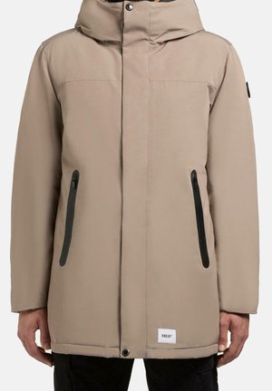 Veste d'hiver beige matelassée avec capuche, deux poches verticales à fermeture éclair noire, fermeture à boutons-pression et fermeture éclair avant, portée avec un pantalon noir.