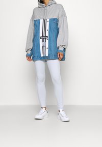 Jeansjacke und graue Kapuzenjacke mit einem weißen und schwarzen gestreiften Vorderteil, versehen mit Logodetails. Kombiniert mit weißen Leggings und Sneakers.