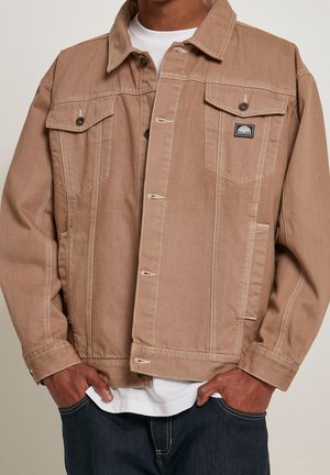 Mann trägt eine beige Knopf-Denimjacke mit Klappentaschen auf der Brust und einem kleinen schwarzen rechteckigen Logo-Patch auf der linken Seite.