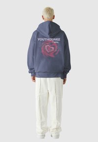 Laimīgā jūras zila kapuce ar grafisku tekstu uz muguras, apvienota ar baltām teksturētām biksēm ar redzamām šuvēm un brīvu piegriezumu.