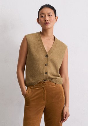 Frau mit dunklem Haar in einem beigen ärmellosen Strick-Button-Top und hoch taillierten braunen Cordhosen, die vor einer weißen Wand steht.