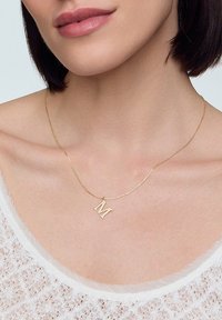 Złoty łańcuszek z wisiorkiem w kształcie dużej litery "M". Chain jest cienki i delikatny, doskonale komponując się z lekką, teksturowaną bluzką.