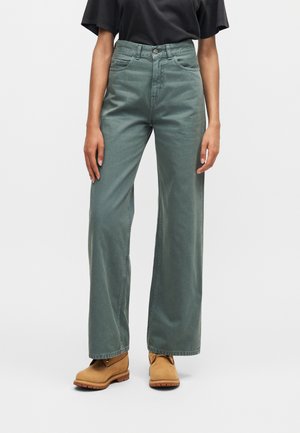 Personne portant un jean vert taille haute à jambes larges, un haut noir à manches courtes et des bottes lacées couleur fauve, debout sur un fond blanc.