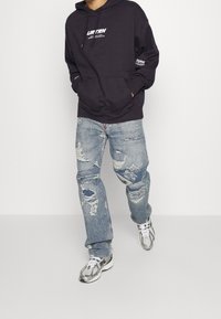 Persona con sudadera oscura, jeans azul desgastados y zapatillas plateadas, caminando contra un fondo blanco liso.