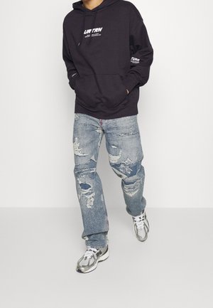 Osoba w ciemnej bluzie z kapturem, zniszczonych niebieskich dżinsach i srebrno-białych sneakersach, idąca na tle białego tła.