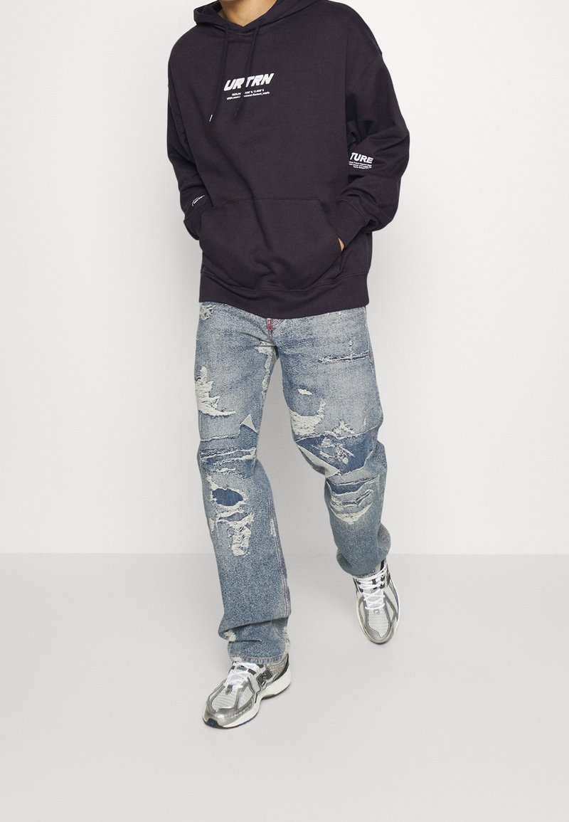 Persona con sudadera oscura, jeans azul desgastados y zapatillas plateadas, caminando contra un fondo blanco liso.