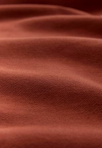 Gros plan sur un tissu lisse et plié avec une texture fine en maille, dans une couleur terre cuite chaude, montrant des jeux subtils de lumière et d'ombre sur la surface.