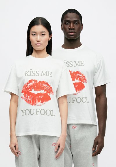 Δύο άτομα φορούν λευκά, φαρδιά t-shirts με ένα κόκκινο αποτύπωμα φιλιού και το κείμενο «ΦΙΛΗΣΕ ΜΕ, ΗΛΙΘΙΕ». Τα t-shirts έχουν χαλαρά μανίκια.