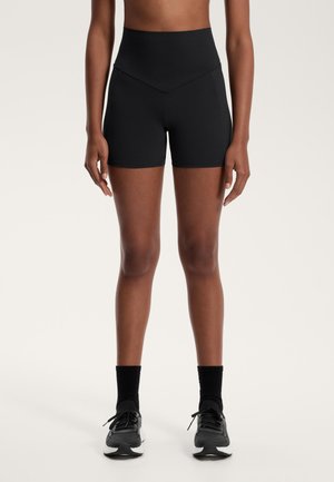 Frau trägt hoch geschnittene schwarze Sportshorts, schwarze Socken und schwarze sowie weiße Laufschuhe, steht mit entspannten Armen an den Seiten.