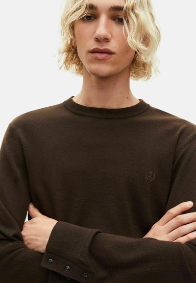 Persona rubia con cabello ondulado que lleva un suéter de punto marrón oscuro con un pequeño logo bordado y detalles de botones en las mangas, con los brazos cruzados.