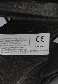 Sorte hjelm med en struktureret overflade, der har ventilationshuller. Indeholder en hvid label med sikkerhedsinformation og certifikationsdetaljer.
