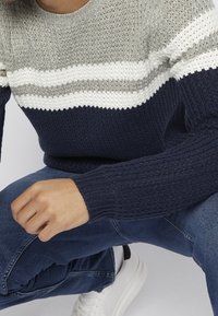 Personne portant un pull en maille gris, blanc et bleu marine, un jean bleu et des baskets blanches, accroupie avec une main posée sur le genou.
