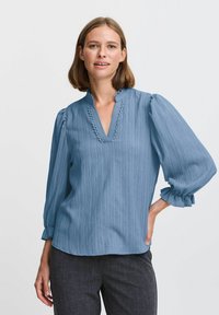 Blusa azzurro chiaro con scollatura a V, bordo arricciato e maniche a sbuffo. Tessuto a trama con sottili righe verticali, abbinata a pantaloni grigio scuro.