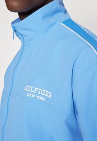 Chaqueta azul claro con cremallera, con cuello alto, logo blanco en el pecho y panel azul texturizado en el hombro. Acabado de tela lisa.