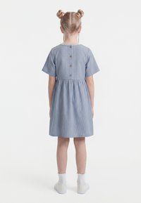 Mergaitė su dviem plaukų kuodeliais dėvi mėlynos ir baltos spalvos dryžuotą trumpomis rankovėmis suknelę su sagutėmis nugaroje, baltas kojines ir baltus sportinius batelius.