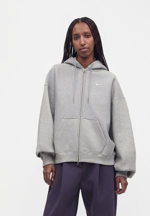 Модел с дълги плитки носи светлосив суичър Nike с цип и тъмнолилави панталони пред обикновен бял фон.