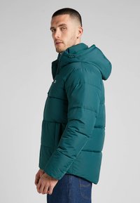 Jasnoniebieska kurtka puchowa z kapturem, posiadająca pikowane elementy, zamek błyskawiczny z przodu oraz regulowane sznurki, noszona z ciemnymi dżinsami.