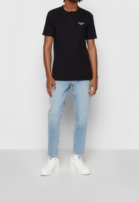 Černé polo tričko s krátkým rukávem a zapínáním na knoflíky, se znakem v bílé barvě. Spárováno se světle modrými slim-fit džíny a bílými teniskami.