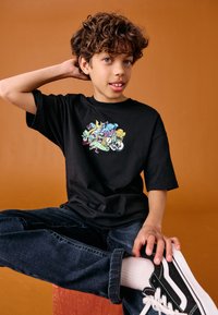Black cotton t-shirt featuring colorful cartoon graphic, paired with dark denim jeans and black Vans sneakers. Warm orange background.