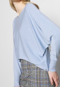 Kvinna som bär en ljusblå tröja med lång ärm och en högmidjad rutig kjol med blå och gröna ränder.