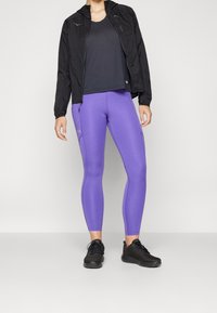 Persona che indossa una giacca nera con zip, una canotta nera, leggings viola e scarpe da atletica nere, in piedi contro uno sfondo chiaro e neutro.