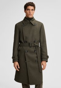 Manteau en mélange de laine vert olive croisé à double boutonnage avec taille ceinturée, grands boutons noirs et col montant, présentant un design mi-long.