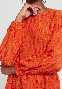 Orangefarbene Bluse mit strukturiertem Stoff und gepufften Ärmeln, die aus transparentem Material mit vertikalen, flauschigen Streifen besteht. Mit einem Rundhalsausschnitt abgerundet.